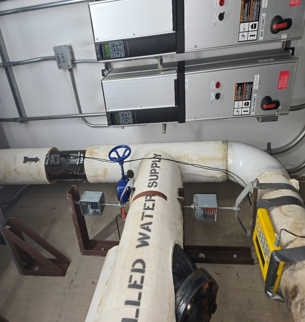 Portaflow PT330 clamp-on ultrasonic flow meter strapped to the chilled water supply main beside pump VFD panels